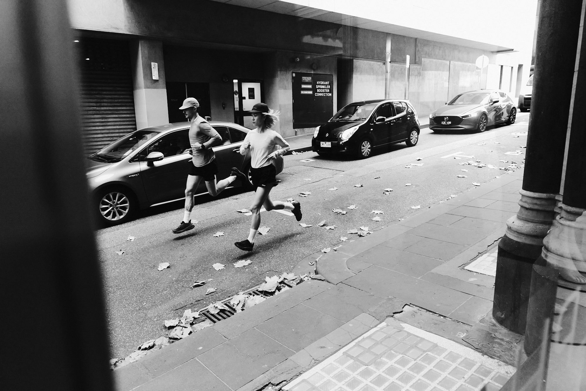 Two people running on a city street with cars parked along the side.