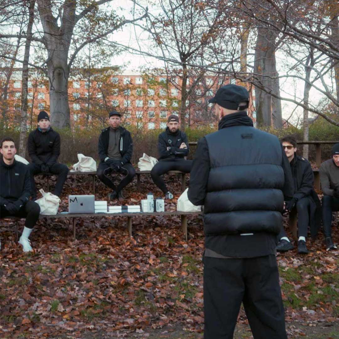Group of people sitting on benches in a park with a person doing an introduction to Metal Health Club by DOXA RUN.